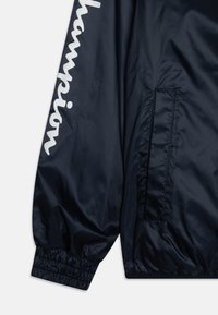 Marinblå vindjacka i glänsande nylon, med elastisk midja och ärmslut, sidoficka och framträdande vit "Champion"-logotyp på ärmen.