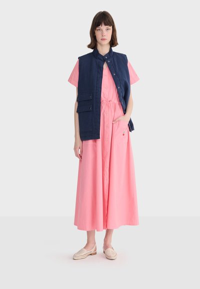 Femme portant une longue robe rose boutonnée à manches courtes et un gilet sans manches bleu marine avec poches à rabat, debout avec des chaussures neutres.