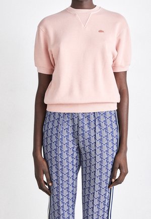 Personne portant un pull léger rose pâle à manches courtes et un pantalon bleu avec un motif géométrique blanc et marine.