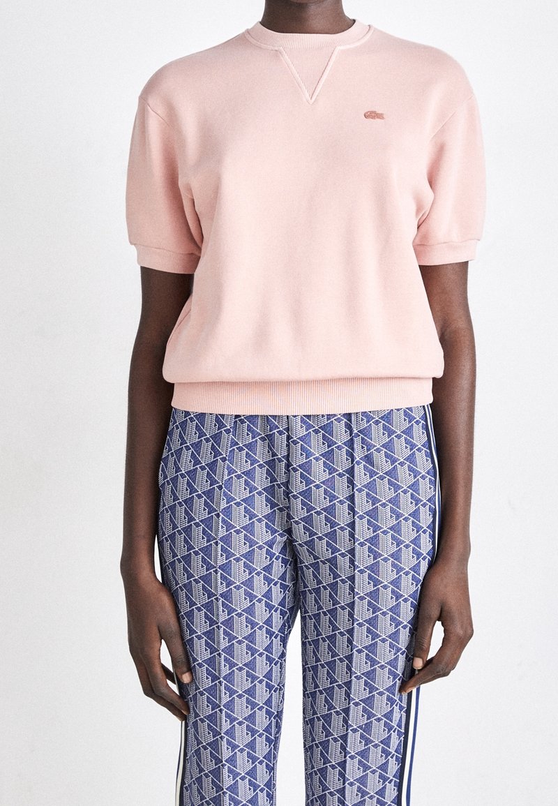 Personne portant un pull léger rose pâle à manches courtes et un pantalon bleu avec un motif géométrique blanc et marine.