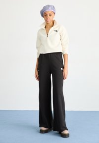 Pullover in fleece crema con zip a metà, pantaloni a gamba larga neri e sandali con piattaforma beige, con una patch logo sui pantaloni.