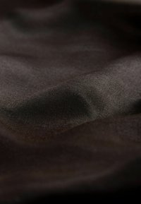 Dunkelbrauner Stoff mit einer glatten, matten Textur. Das Material wirkt weich, leicht zerknittert und zeigt subtile Farbtonvariationen.