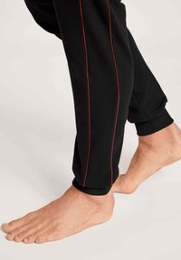Un pantalon de survêtement noir à coupe ajustée avec des rayures rouges sur les côtés. Les poignets sont côtelés et le tissu semble doux. Un pied nu est visible.