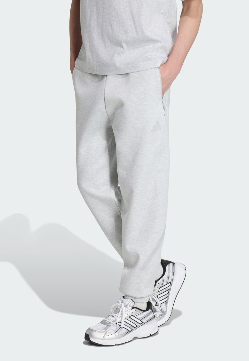 Personne portant un pantalon de survêtement gris clair et un haut assorti avec des baskets Adidas blanches et noires, debout les mains dans les poches.