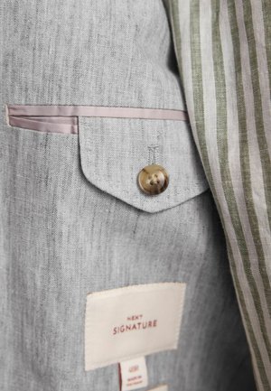 Blazer de lino gris con un bolsillo en el pecho con un botón marrón, complementado por un forro interior a rayas blancas y verdes y etiquetas de cuidado.