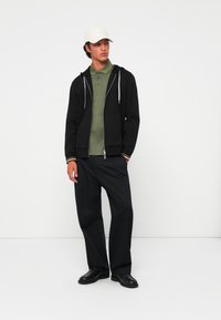 Felpa nera con zip e cordini grigi sopra una polo verde testurizzata, abbinata a pantaloni neri larghi e scarpe nere. Cappellino bianco indossato.