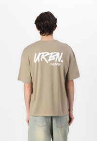 Tricou din bumbac bej, cu croială lejeră, având un mare text grafic alb "URBN." și "CULTURE" pe spate, textură netedă și decolteu rotund.