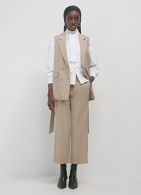 Frau in beigem ärmellosem Blazer und Hose, weißer Bluse mit hohem Kragen und weiten Manschettenärmel, schwarzem Kopftuch und schwarzen Ankle Boots.