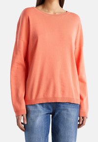 Korálový svetr s kulatým výstřihem, spuštěnými rameny, žebrovanými manžetami a lemem. Materiál vypadá jemně a má hladký povrch. Spárovaný s modrými džíny.