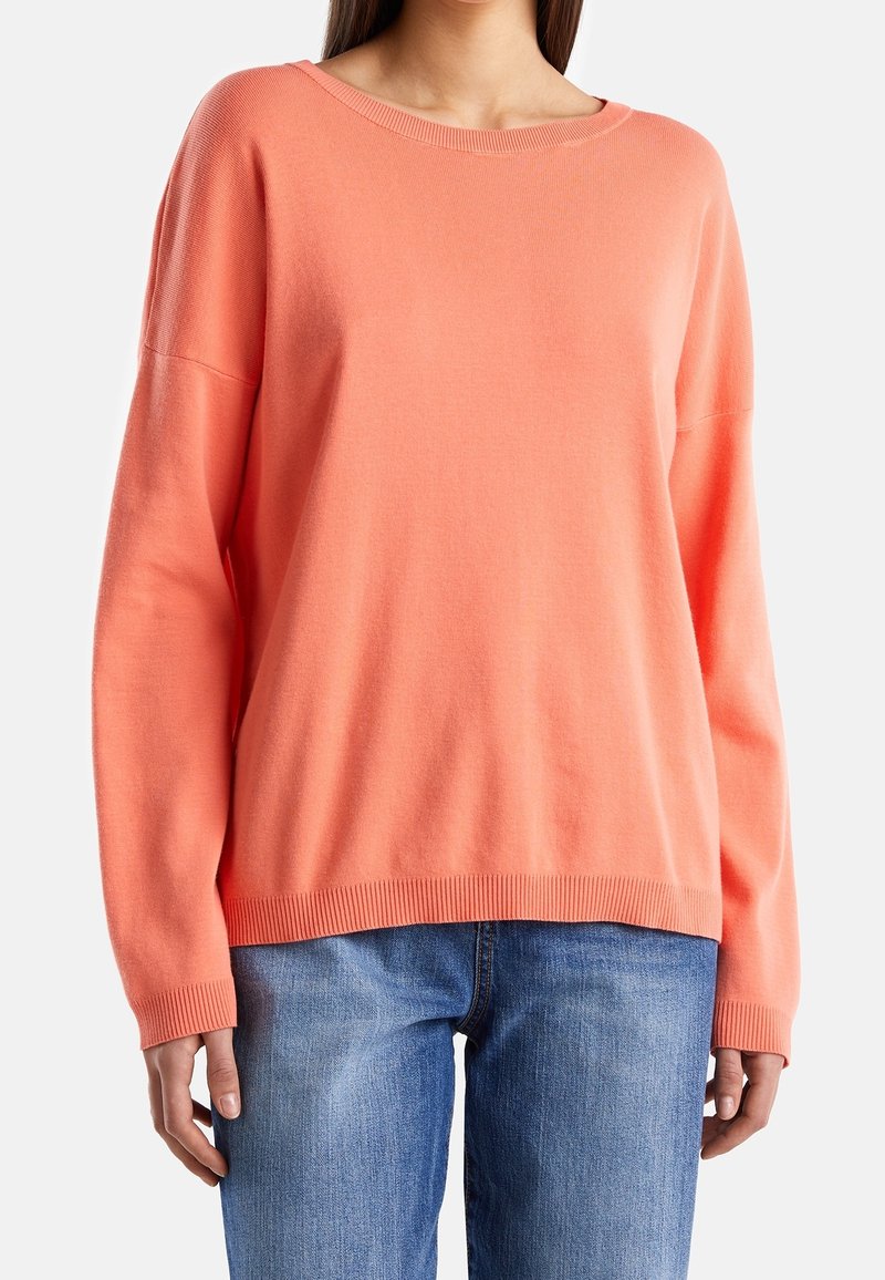 Korálový svetr s kulatým výstřihem, spuštěnými rameny, žebrovanými manžetami a lemem. Materiál vypadá jemně a má hladký povrch. Spárovaný s modrými džíny.