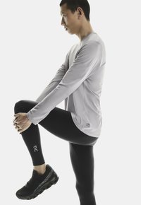 Grå långärmad tröja, svarta åtsittande leggings och svarta sportskor med en strukturerad sula. Modell i en sidosträckningsposition.