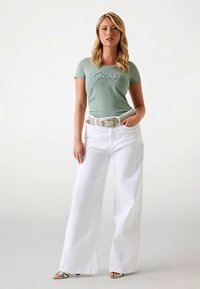T-shirt verde chiaro aderente con logo "Guess", abbinata a pantaloni bianchi a vita alta e gamba larga. Cintura strutturata e sandali metallizzati con cinturini.