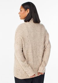 Beige gestrickter Pullover mit hohem Kragen, gerippten Bündchen und schwarzen Sprenkeln auf dem Stoff. Hat eine lockere Passform und seitliche Schlitze.
