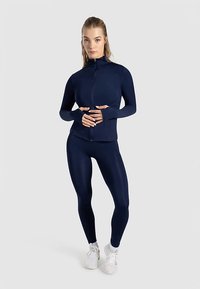 Marinblå atletisk set med en figursydd zip-up jacka med hög krage och tumhål, tillsammans med matchande leggings i en slät textur.