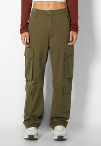 Olijfgroene cargobroek van katoen, met meerdere zijzakken en een ontspannen pasvorm. Draagt witte sneakers.