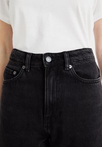 Osoba w czarnych dżinsach z wysokim stanem i prostym białym t-shircie, skupiona na okolicy pasa z zapiętym guzikiem.