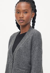 Grå cardigan laget av teksturert stoff, med v-hals, to knappelukkinger og rhinestones i kanten foran.