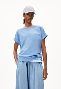 Himmelblaues Baumwoll-T-Shirt mit aufgerollten Ärmeln, das ein kleines Logo auf der Brust trägt; kombiniert mit einem hellen Jeansrock und einer grauen Kappe.