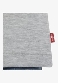 Camiseta de algodón gris con una textura suave, que presenta una etiqueta de marca roja visible en el dobladillo, expuesta sobre un fondo de mezclilla azul.