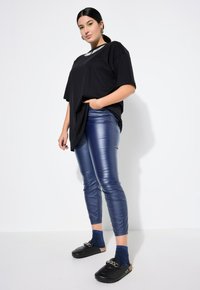 Zwarte oversized t-shirt, glanzende blauwe nepleer leggings, zwarte klompen met kettingdetails en blauwe sprankelende sokken op een simpele achtergrond.