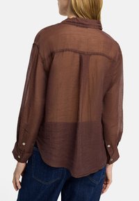 Brąsza półprzezroczysta koszula zapinana na guziki z długimi rękawami i zaokrąglonym dołem, z delikatną teksturą i detalem kołnierza, w połączeniu z niebieskimi dżinsami.