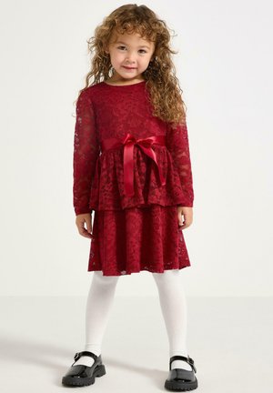 Abito rosso in pizzo con maniche lunghe e design a strati, caratterizzato da una cintura in satin. Collant bianchi e scarpe Mary Jane nere e lucide completano l'outfit.