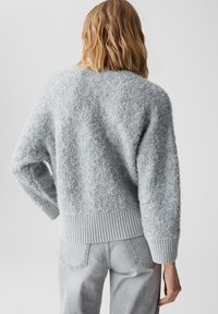 Person mit schulterlangen blonden Haaren, die einen hellgrauen strukturierten Pullover und passende hellgraue Hosen trägt und mit dem Rücken zur Kamera steht.