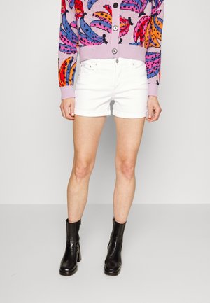 Pantalones cortos de mezclilla blanca con dobladillos enrollados, combinados con botas de tobillo negras con tacones. Un suéter de colores brillantes presenta un patrón de plátanos y botones.