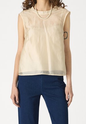 Vrouw draagt een mouwloze doorzichtige beige top, donkerblauwe broek, parelketting en zichtbare zwarte ovale tatoeage op de bovenste linkerarm.