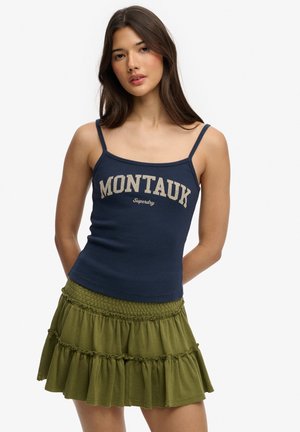 Femme portant un débardeur bleu marine à bretelles fines avec le texte "MONTAUK" et une mini-jupe à volant olive, debout devant un fond blanc.