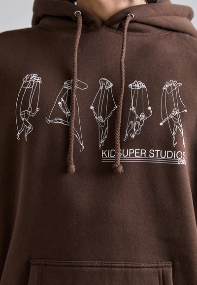 Brown hoodie featuring white line-drawn figures suspended by strings. The design includes a logo that reads "KID SUPER STUDIOS 2022."
