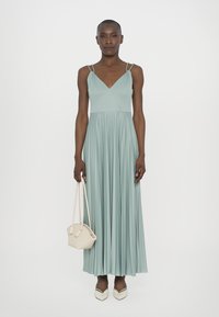 Robe maxi plissée vert menthe avec un décolleté en V, des straps fins, associée à un petit sac tissé et des escarpins blancs pointus.