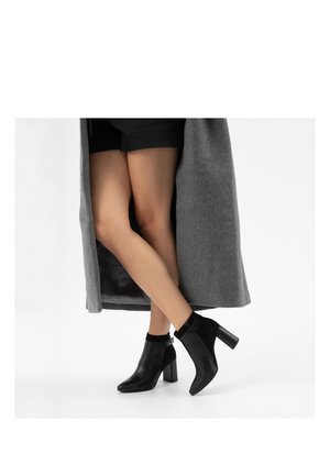 Žena oblečená v černých kotníkových botách na hranatém podpatku, černých šortkách a dlouhém šedém kabátě, stojící se zkříženýma nohama na bílém pozadí.