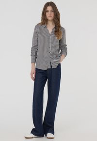 Camicia a righe bianco e nero con maniche lunghe, abbinata a pantaloni larghi in denim scuro e sneakers bianche.