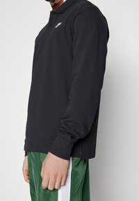 Mężczyzna w czarnej koszulce Nike z długim rękawem i zielonych spodniach sportowych z białym paskiem, stojący na tle jasnym.