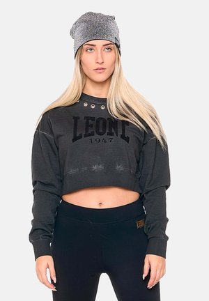 Vrouw met lang blond haar die een grijze muts draagt, een cropped zwarte sweatshirt met "LEONE 1947" en zwarte tight tegen een effen achtergrond.
