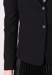 Černý blazer se třemi knoflíky, nošený přes skladanou černou sukni. Zaměření na detaily rukávu a saka, které ukazují elegantní, profesionální vzhled.