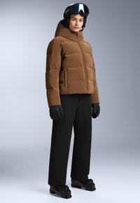 Hnědá péřová bunda s kapucí, se zipem vpředu, v kombinaci s černými lyžařskými kalhotami a rukavicemi. Nosit s černými lyžařskými botami s přezkami.