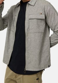 Homme portant une veste beige zippée par-dessus une chemise noire et un pantalon kaki, debout avec un bras plié et la main près du visage.
