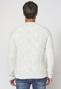 Vit stickad tröja som har ett strukturerat mönster med diamants former, ribbade ärmslut och nederkant, visas framifrån mot en enkel bakgrund.