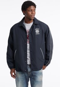 Marineblauer Windbreaker mit weißen Streifen entlang der Ärmel, Druckknöpfen und einem Logo auf der Brust; gefüttert mit elastischen Bündchen und Saum.
