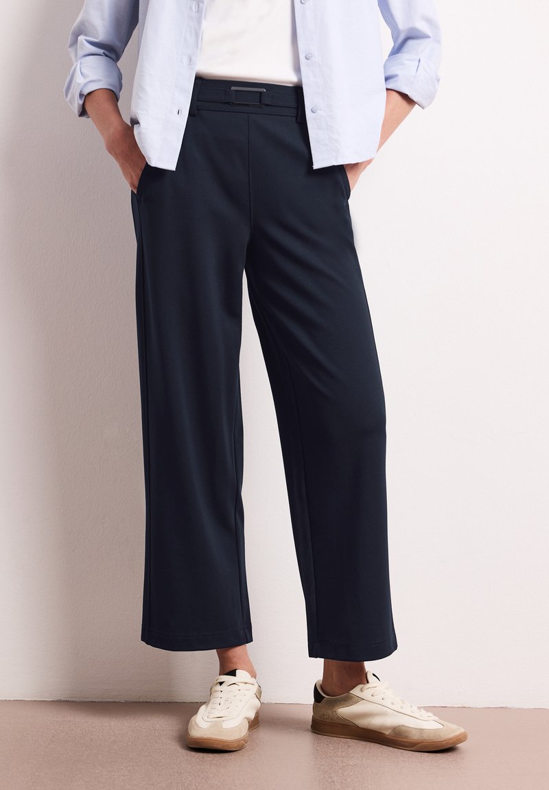 Personne portant un pantalon noir court, des baskets blanches et une chemise bleu clair boutonnée, les mains dans les poches.