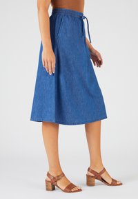 Falda midi de mezclilla con cintura elástica y cordón. Presenta bolsillos laterales y una silueta fluida. Combinada con sandalias de tacón marrones.