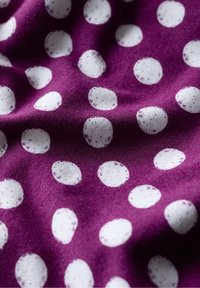 Fioletowa tkanina z rozsianym wzorem nieregularnych białych kropek. Tekstura wydaje się gładka i miękka z drobnymi wariacjami w kształcie kropek.