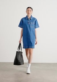 Denim skjortekjole med korte ermer, lyseblå farge, brystlomme og frynset kant. Kombinert med en svart tote bag og hvite sneakers.