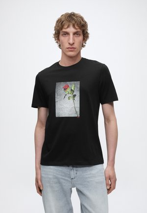 Giovane uomo che indossa una maglietta nera con una grafica di rosa rossa sul davanti, abbinata a jeans azzurri, in piedi davanti a uno sfondo bianco semplice.