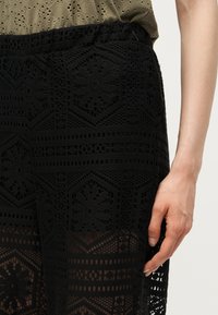 Gros plan d’une personne portant un pantalon noir à motif crochet et un haut vert olive, une main détendue le long du corps.