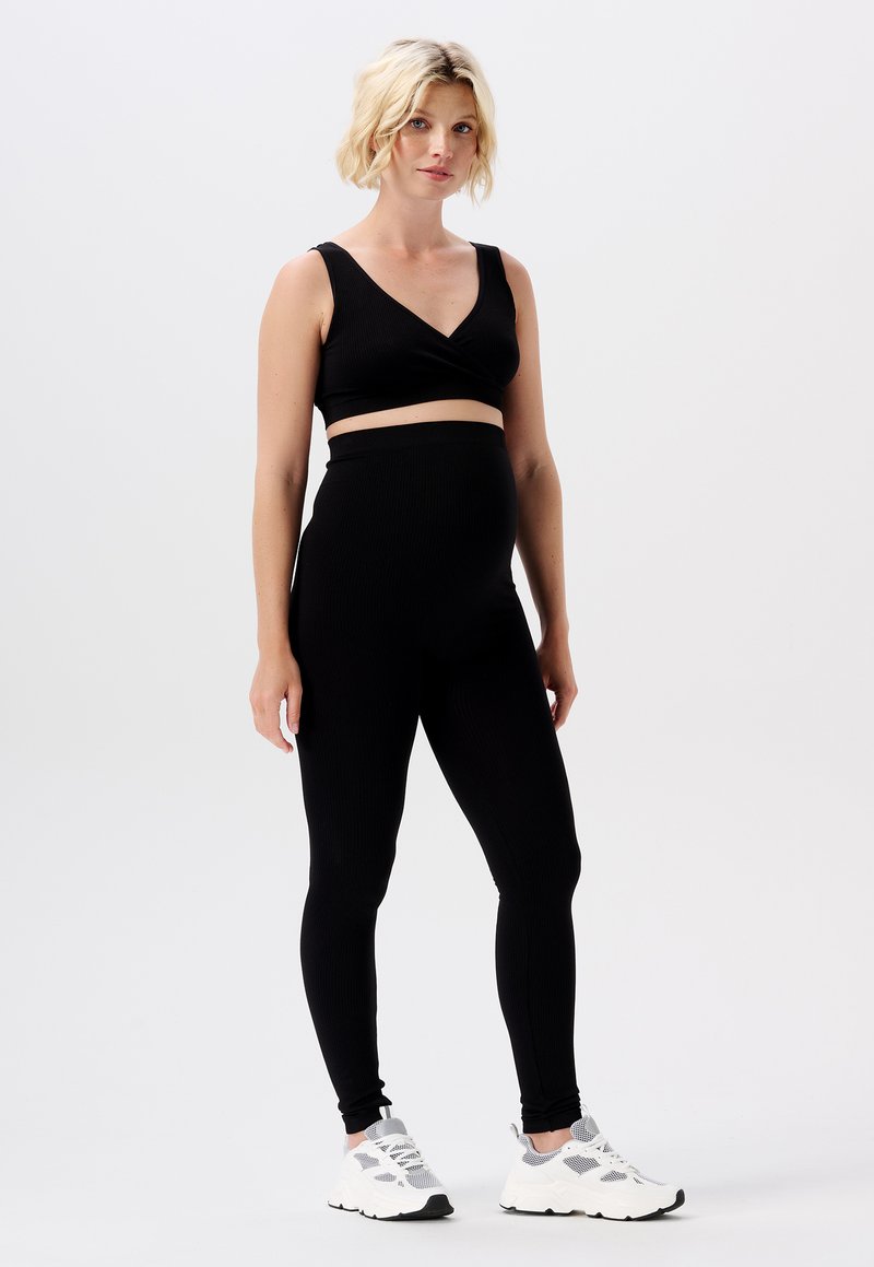 Crop top de maternité noir à côtes avec décolleté en V associé à un legging noir taille haute. Modèle portant des baskets blanches avec des accents gris.