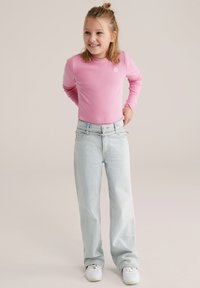 Cămașă roz cu mâneci lungi, cu dungi, asortată cu blugi largi, de culoare albastru deschis. Figura stă cu mâinile pe șold, purtând adidași albi.