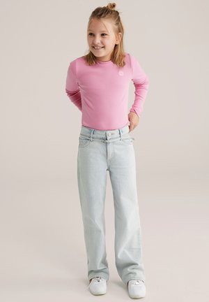 Ružové dlhé tričko s rebrovaným vzorom v kombinácii s voľnými svetlomodrými rifľami. Postava stojí s rukami v bok a má na nohách biele tenisky.